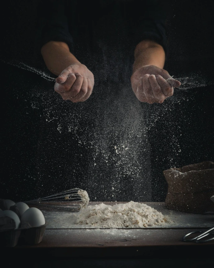 person pouring flour on table beside eggs and whisk